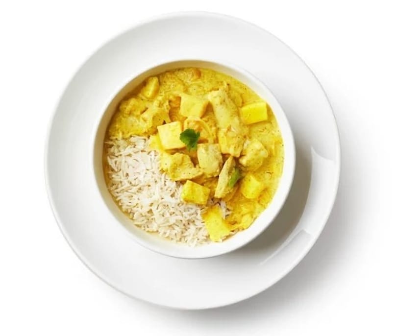 Overhead view of a white bowl filled with yellow curry, rice, and cilantro on a white plate.
