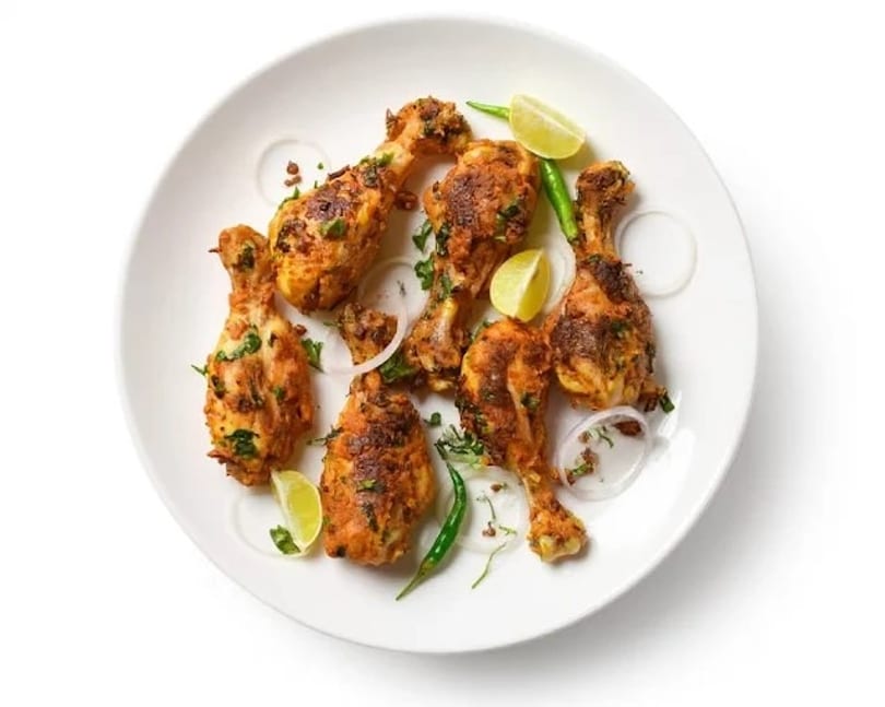 Six chicken drumsticks with herbs, lime wedges, green chilies, and onion rings on a white plate.