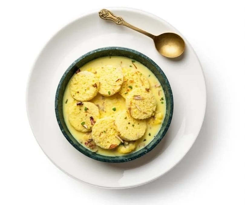 Rasmalai, an Indian dessert, in a dark bowl with a golden spoon on a white plate.